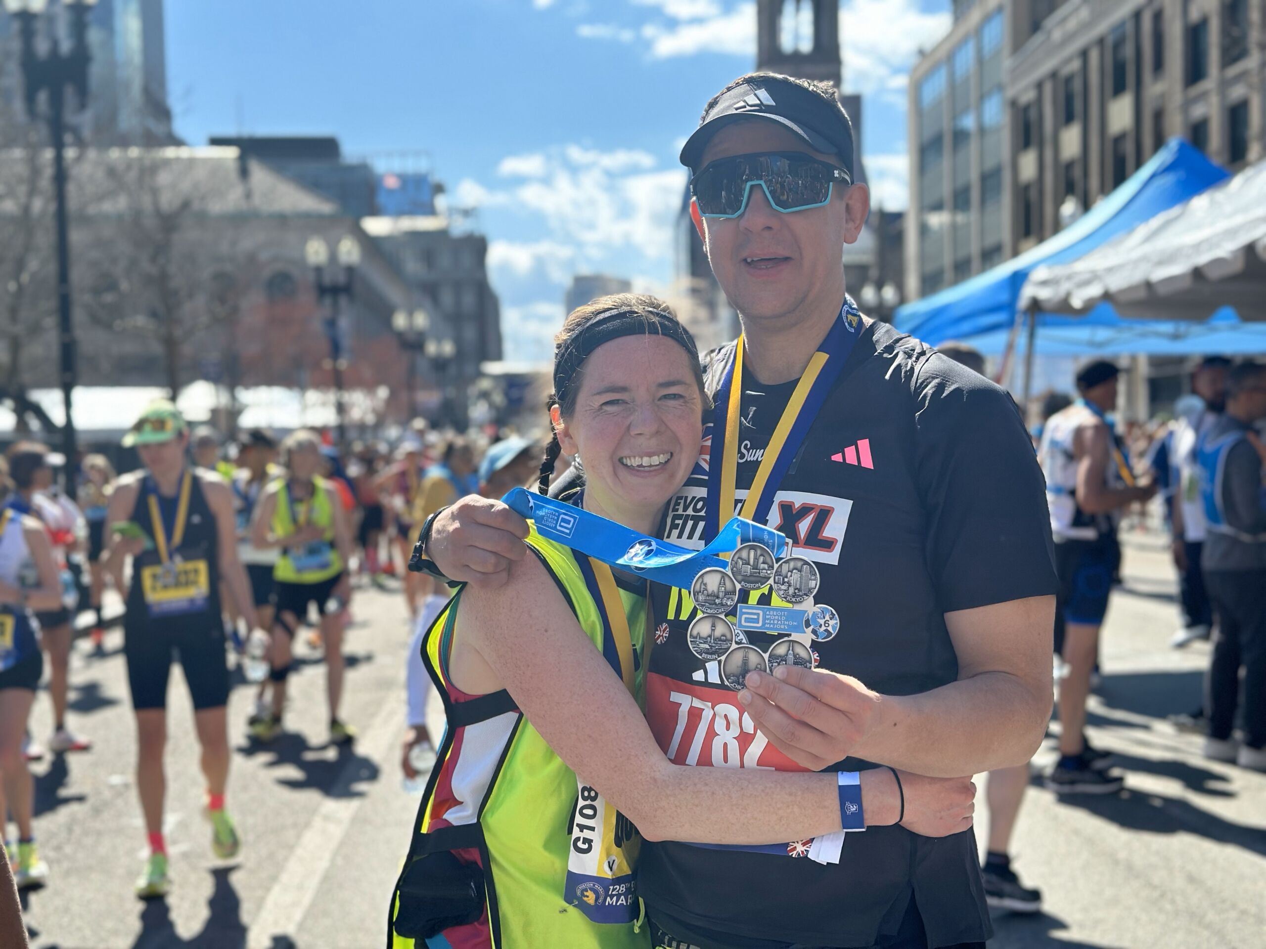 Two runners, one blind and one guide, share a hug at the finish line of the Boston Marathon. One of them holds a medal commemorating the completion of all 6 major marathons.