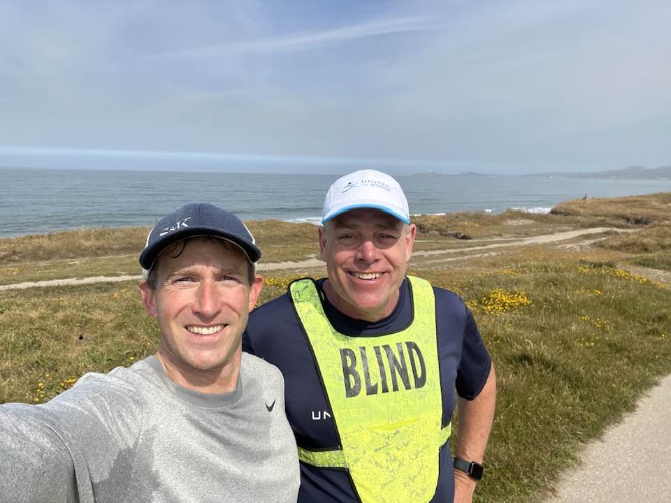 UIS Founder Richard Hunter and guide Chris smile in front of the ocean after a run together. Richard is rocking his United in Stride gear and “Blind” vest.