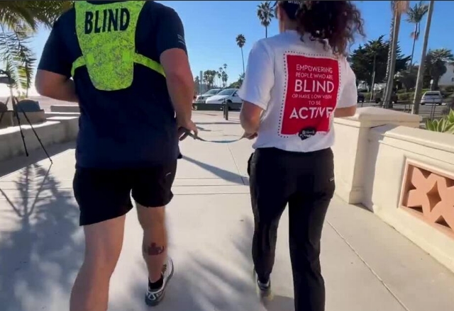 Two runners, holding a tether while running in the sun. One of them is wearing dark clothes and a yellow "Blind" vest, and the other is wearing a white and black pants.