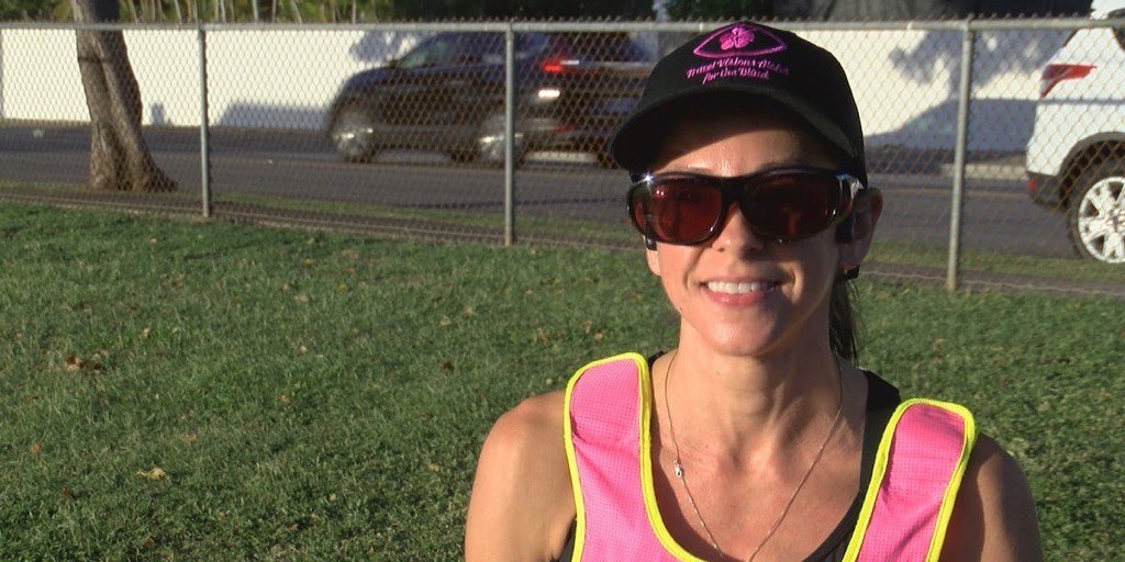 A woman in a hat, sunglasses, and a pink running vest smiling in a grassy area