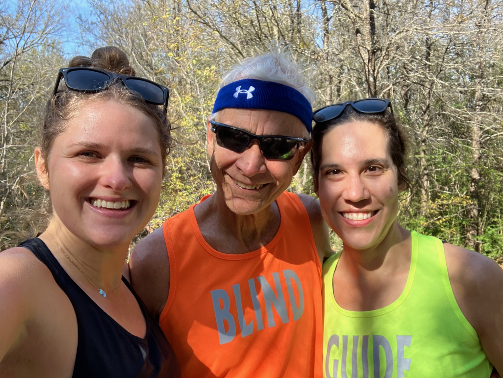 Three runners one wearing a black tank top and sunglesses on her head, one wearing an orange shirt with sunglasses and a tight hat, and one with a bright yellow tank top, all smiling.